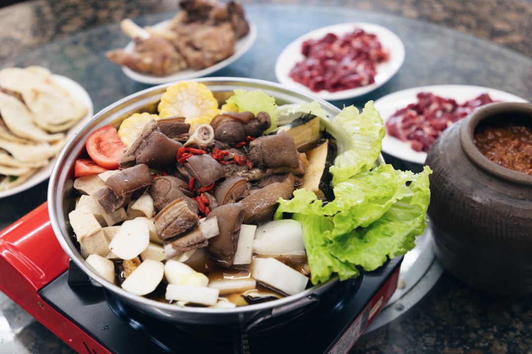 一鍋豐盛的羊肉爐在卡式爐上烹煮，近拍鍋中滿是帶皮羊肉塊、豆腐、玉米與番茄，周圍環繞著生肉片、配菜與沾醬陶甕