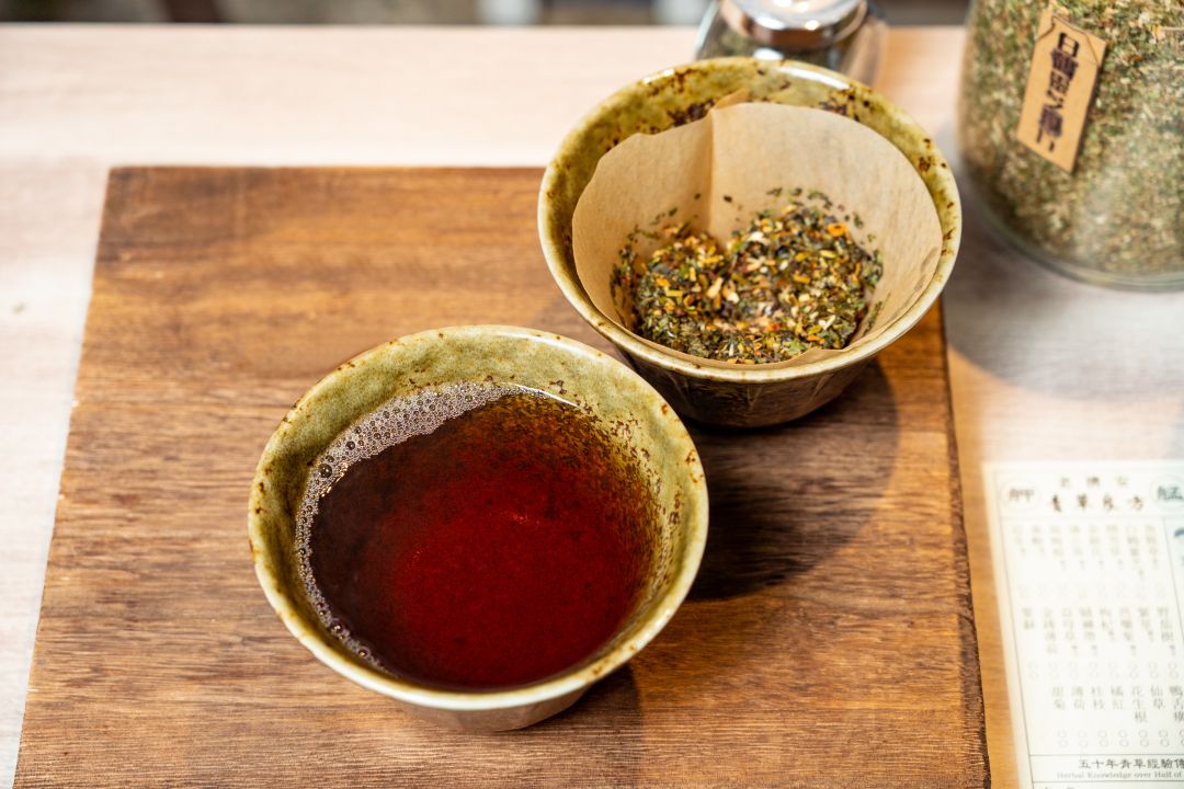 一杯剛泡好的深褐色青草茶盛裝在陶瓷杯中,旁邊是放有乾燥藥草的濾杯,一同置於木製托盤上