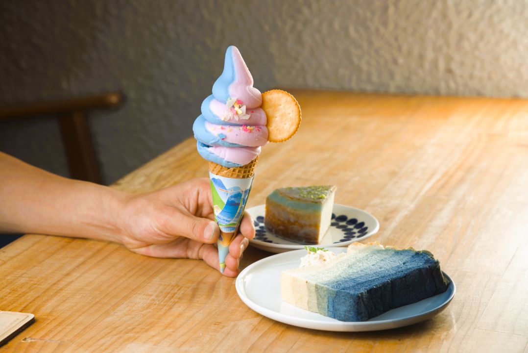 手持藍粉雙色霜淇淋甜筒,木桌上擺放著藍色漸層生吐司與圓形起司蛋糕等特色甜點