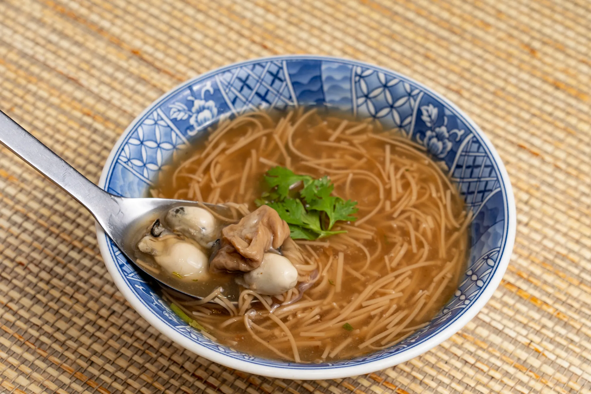 特寫鏡頭展示台灣人氣小吃大腸蚵仔麵線，湯匙舀起肥美蚵仔與滷透的大腸段，搭配滑順的紅麵線與香菜，料多實在。