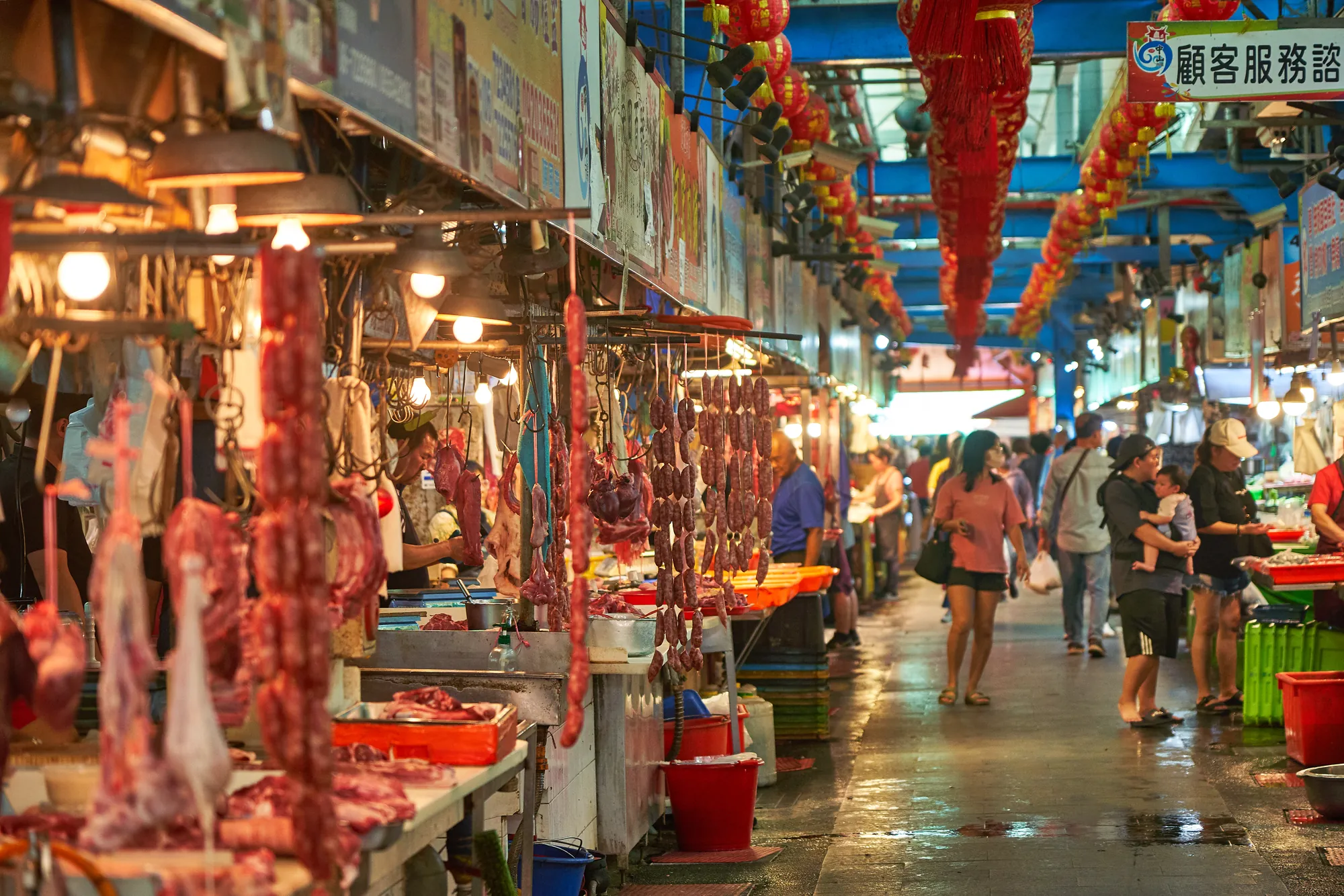 台灣傳統市場內熱鬧的走道，上方掛滿紅色燈籠，攤販懸掛新鮮肉品與手工香腸，顧客穿梭其中，展現道地的庶民生活文化。