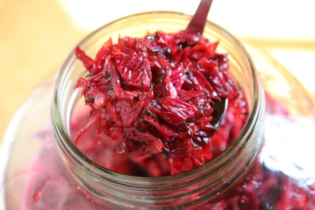 一支金屬湯匙正從玻璃罐中舀出醃漬過的深紅色洛神花（玫瑰茄）果萼特寫