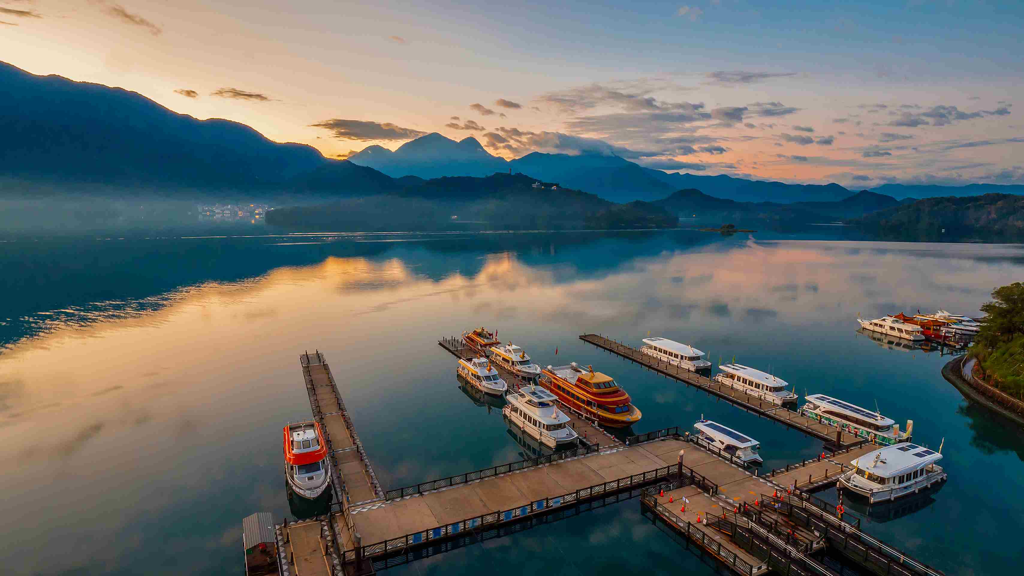 台灣日月潭著名的清晨曙光美景，雲霧繚繞的湖面映照著金色陽光與層疊山巒，碼頭邊整齊停泊著觀光遊艇，呈現靜謐的湖光山色。