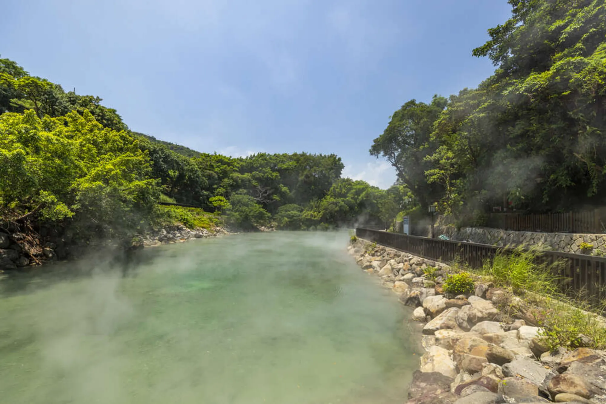 北投地熱谷（鬼湖）的青綠色硫磺泉水冒著滾滾蒸氣，周圍有綠意盎然的樹木與遊客觀賞步道