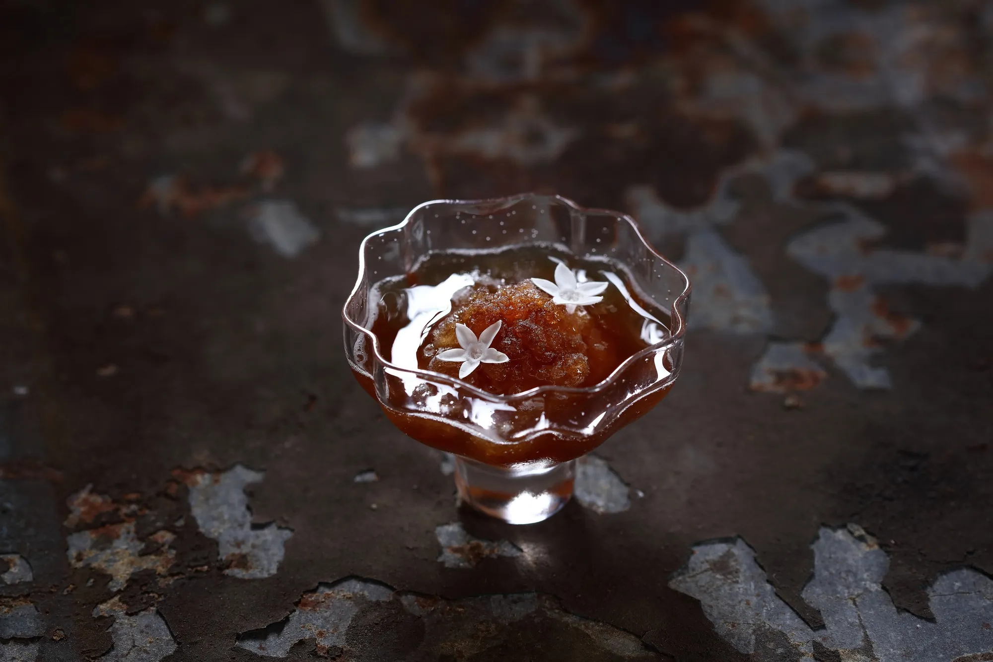 盛裝在精緻玻璃高腳杯中的琥珀色鑽石冰沙，頂端點綴著白色食用花，背景為深色斑駁紋理，展現高端甜點的優雅質感。