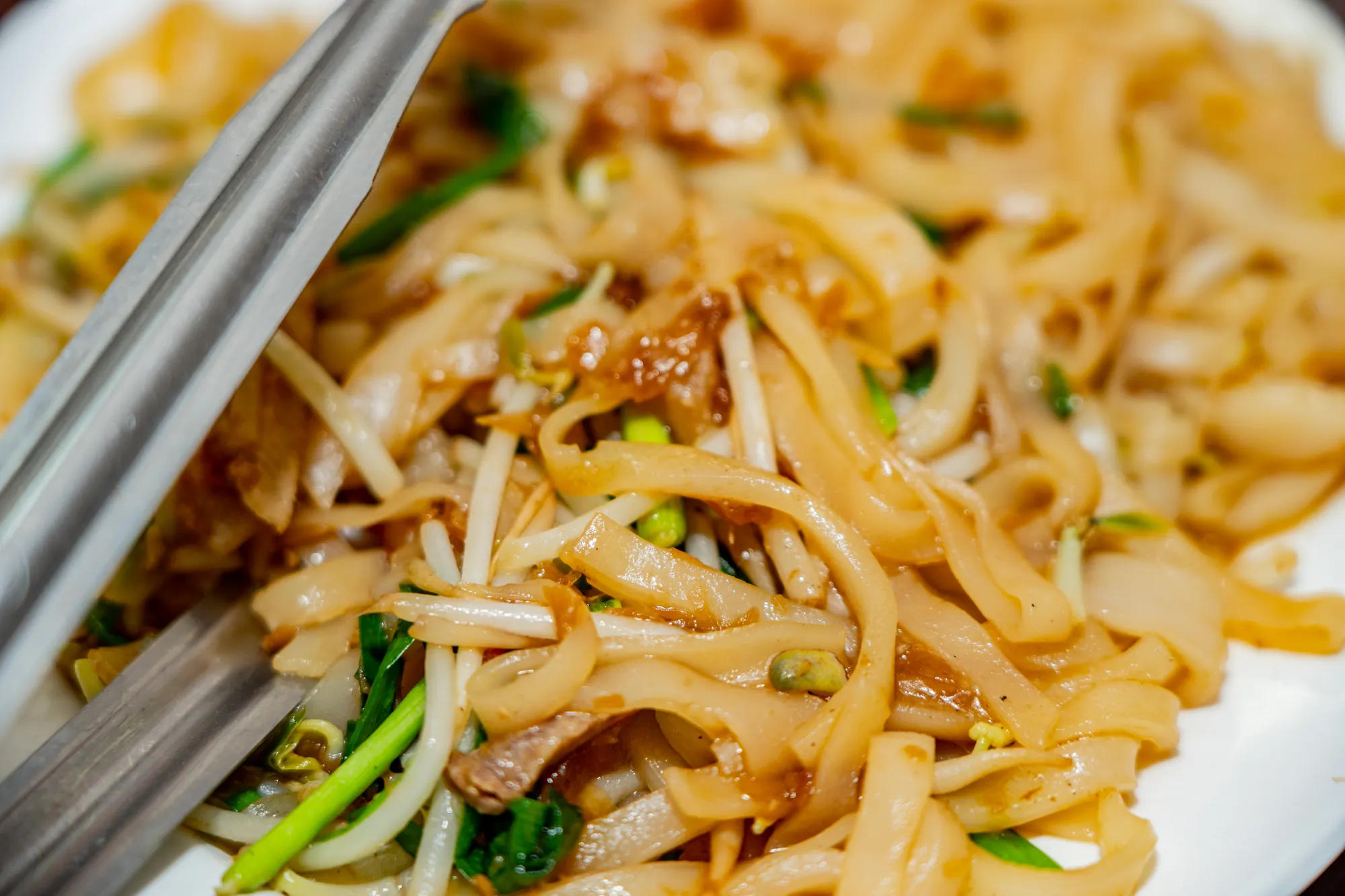 油亮誘人的客家炒粄條特寫，寬扁米麵條吸附醬汁，搭配清脆豆芽與韭菜，盤緣附有夾子，展示台灣道地傳統小吃的美味細節