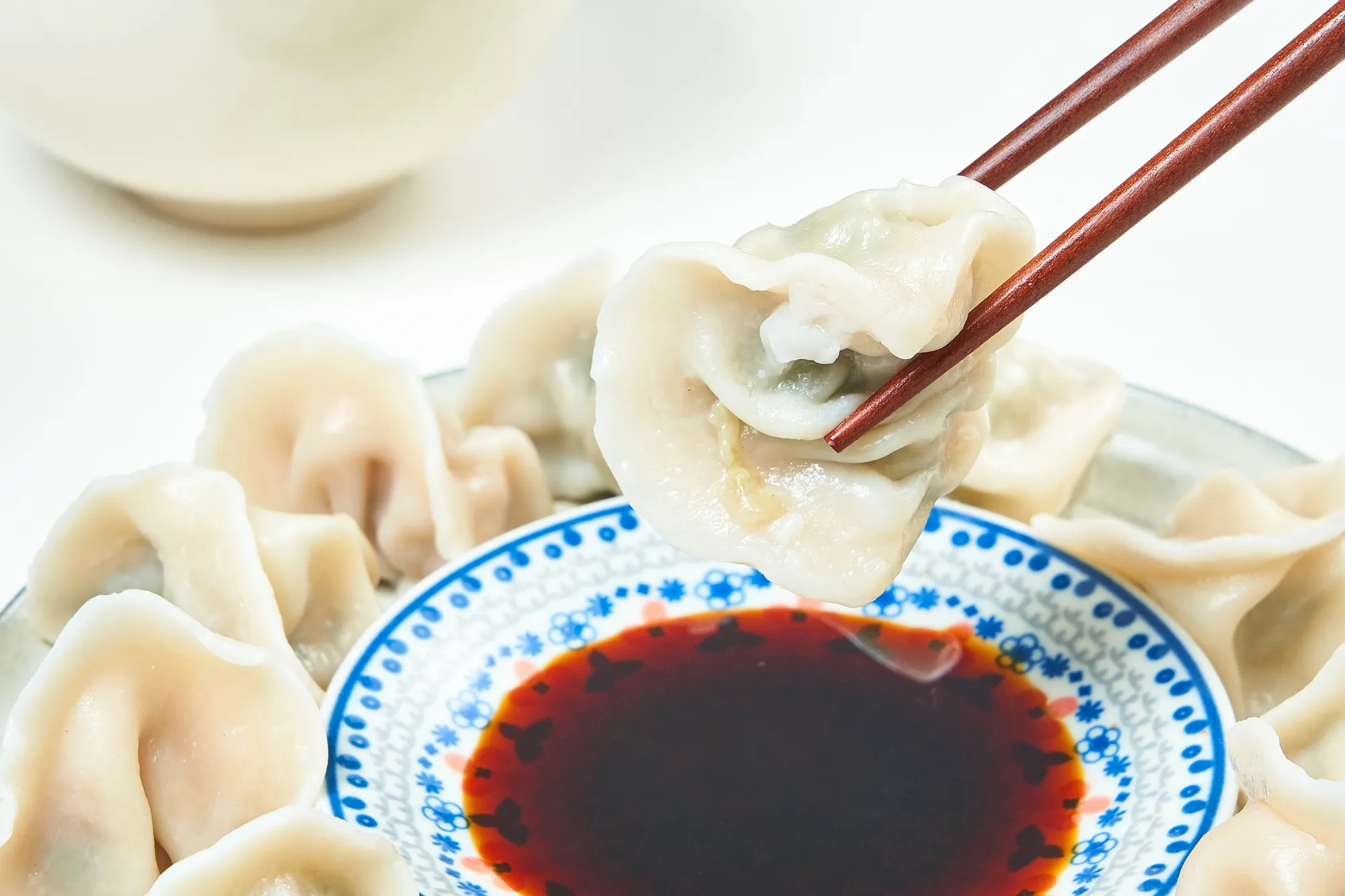 木筷夾起一顆飽滿的韭菜豬肉水餃，背景為裝滿水餃的盤子與一碟深色醬油沾醬