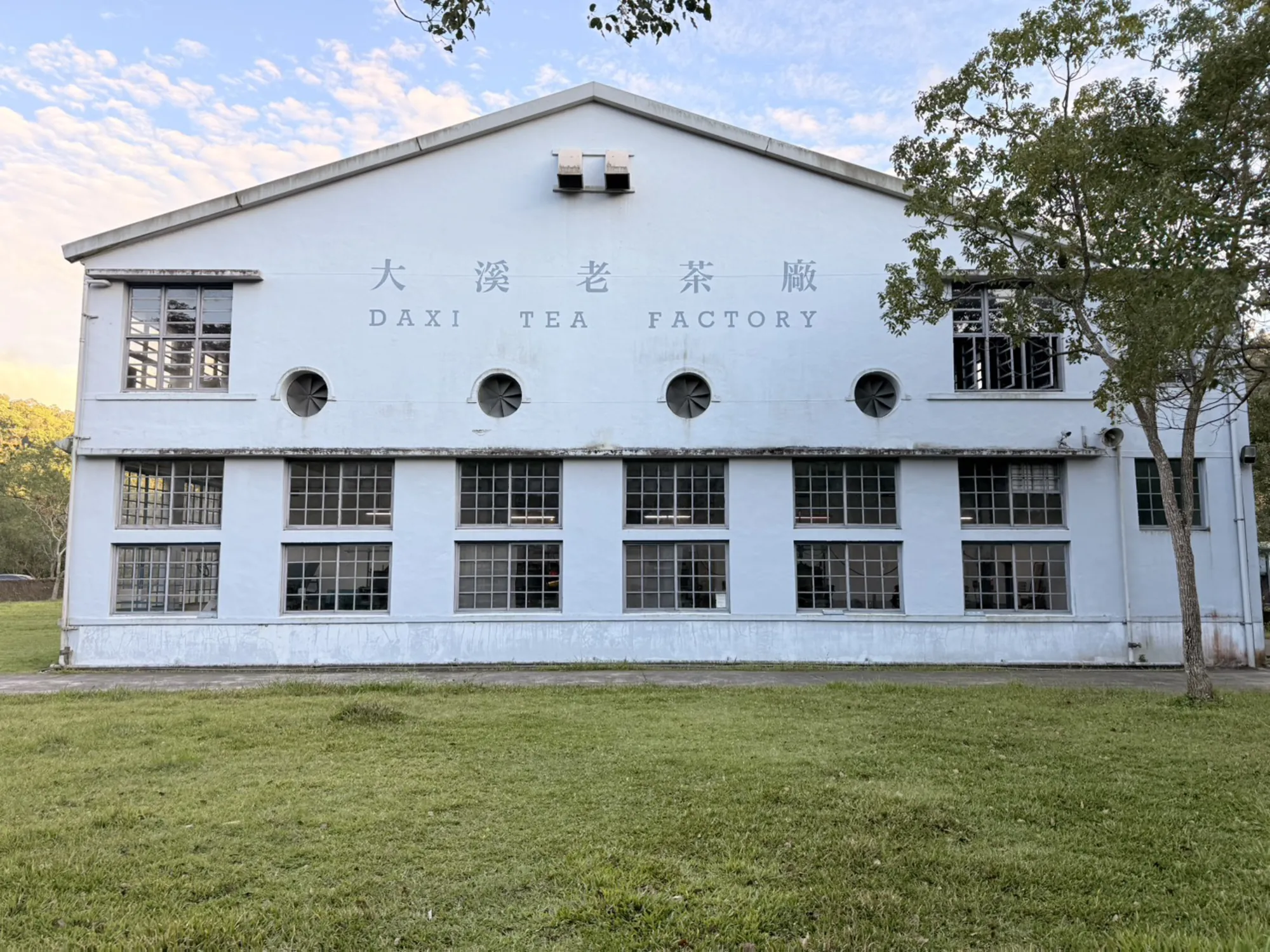 桃園大溪老茶廠白色歷史建築外觀，藍天綠地襯托雙層格窗與中央排氣孔。