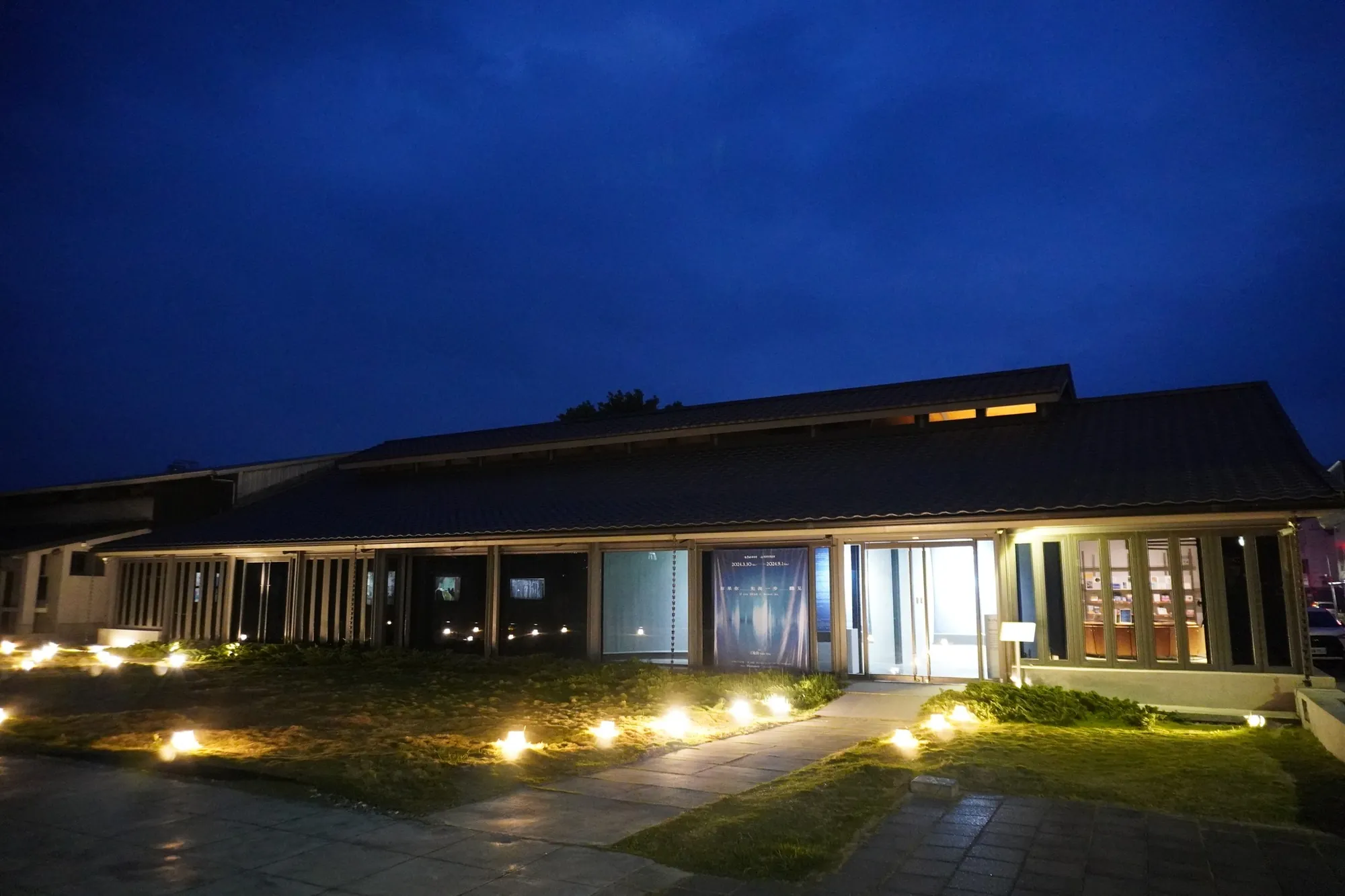 夜晚的台東池上穀倉藝術館外觀，老建築改建的木造建築散發暖光，草地錯落點狀燈飾。展現從米倉轉型為農村藝術展演空間的土地美學。