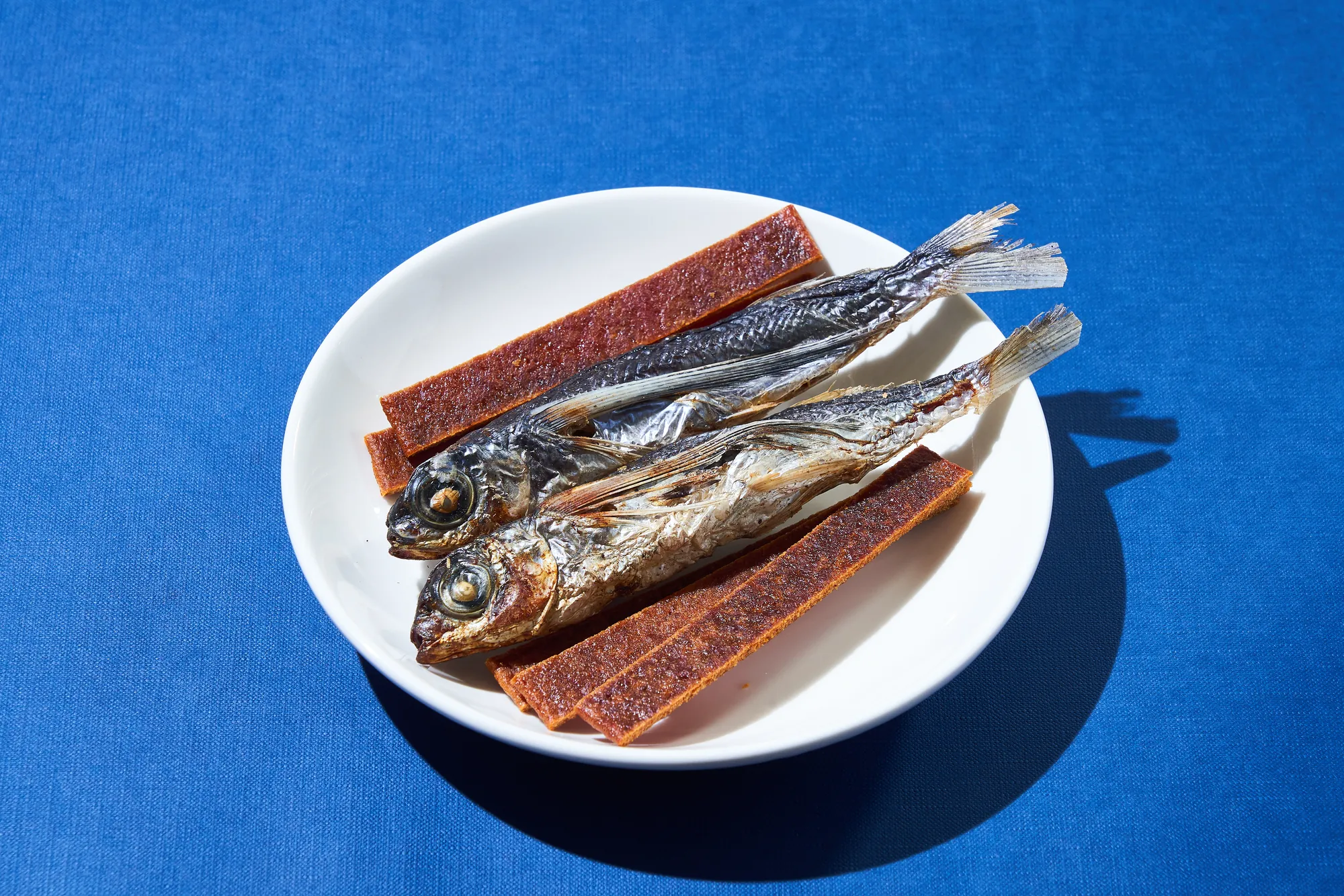 白色圓盤上擺放的兩條煙燻飛魚乾與紅褐色魚肉條，背景為深藍色布面，展現蘭嶼達悟族傳統食魚文化。