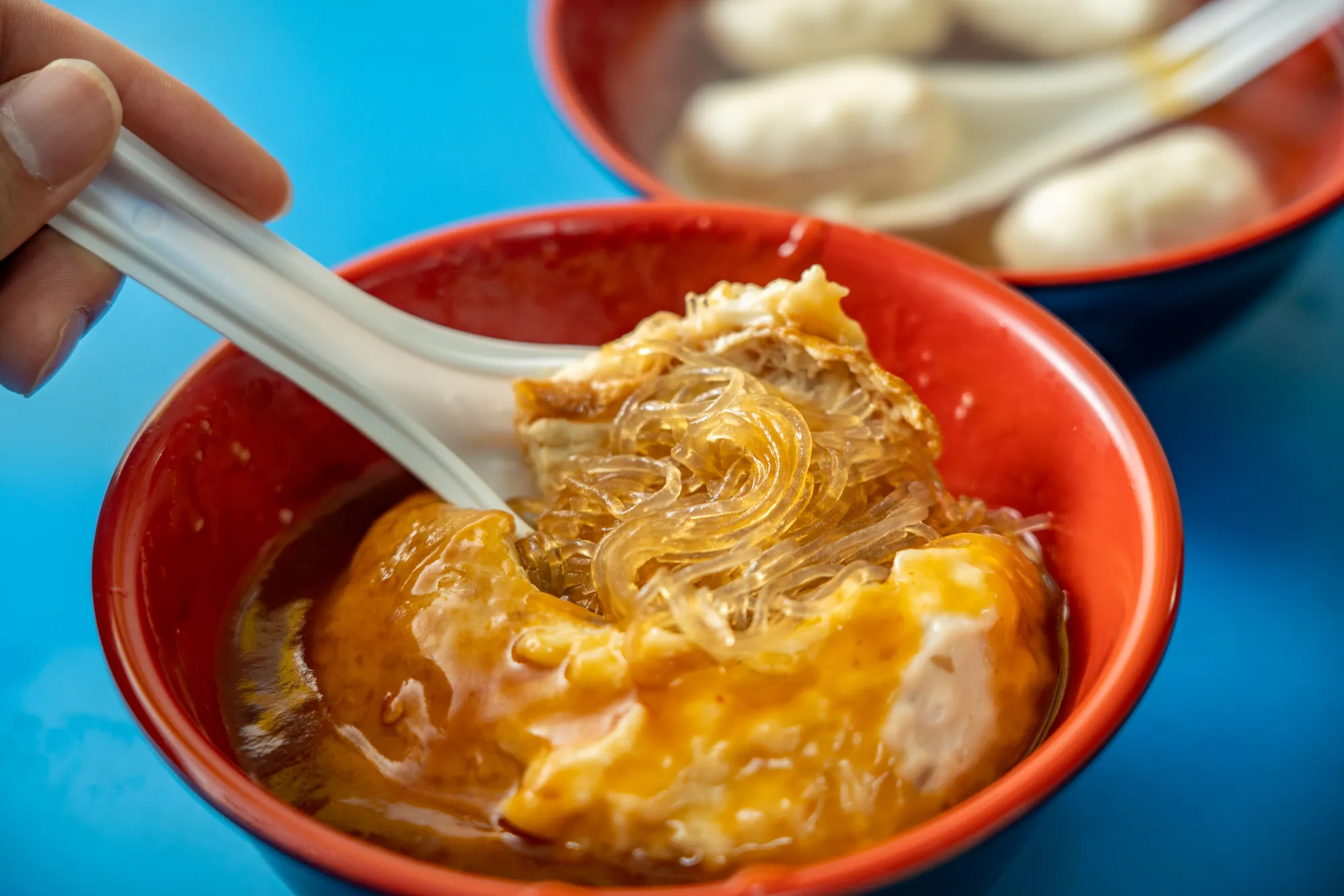 淡水名產阿給特寫，切開的油豆腐露出吸滿醬汁的冬粉，上方淋有甜辣醬，背景為魚丸湯。