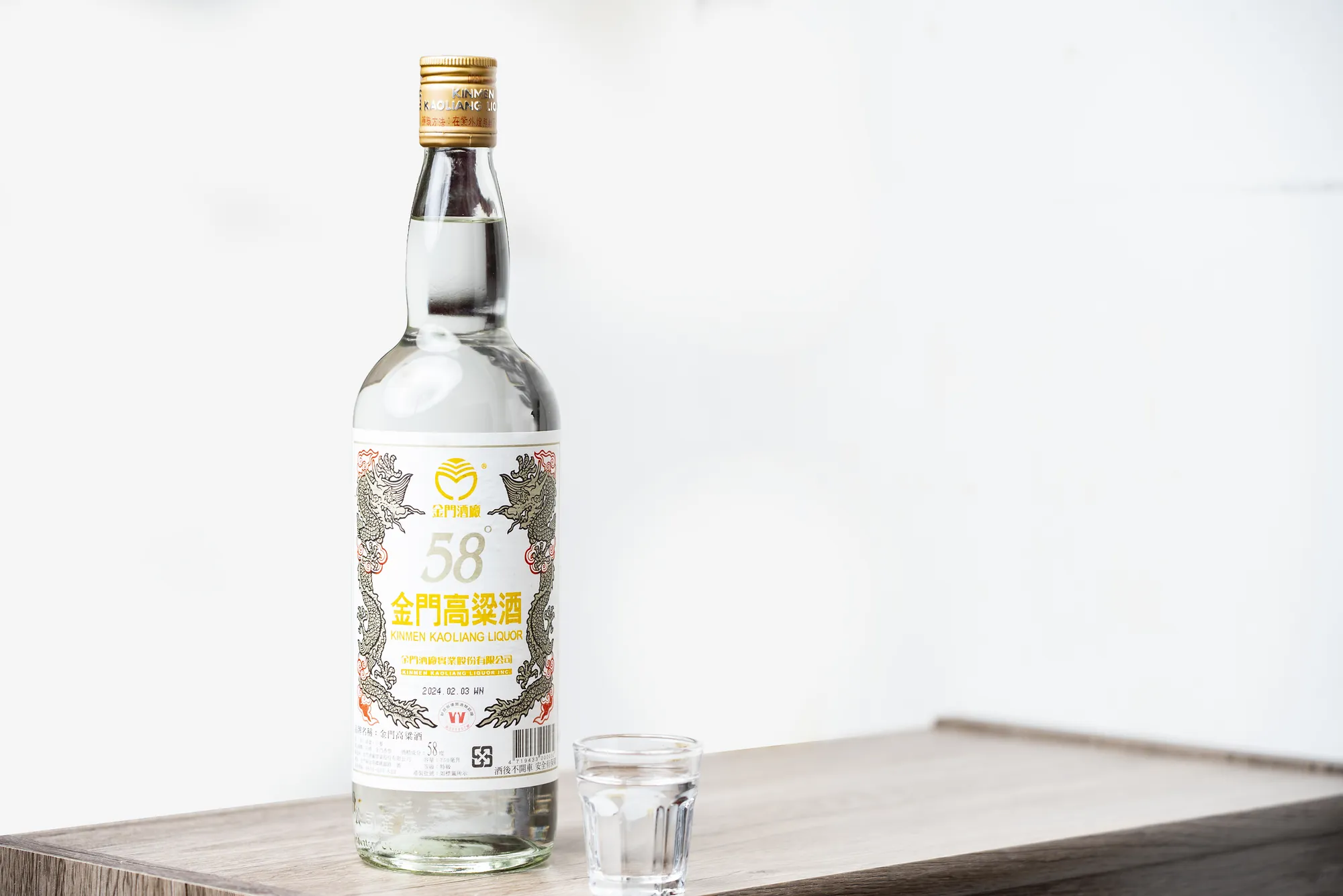 經典 58 度金門高粱酒瓶與小烈酒杯，放置於木質檯面，白色簡約背景。