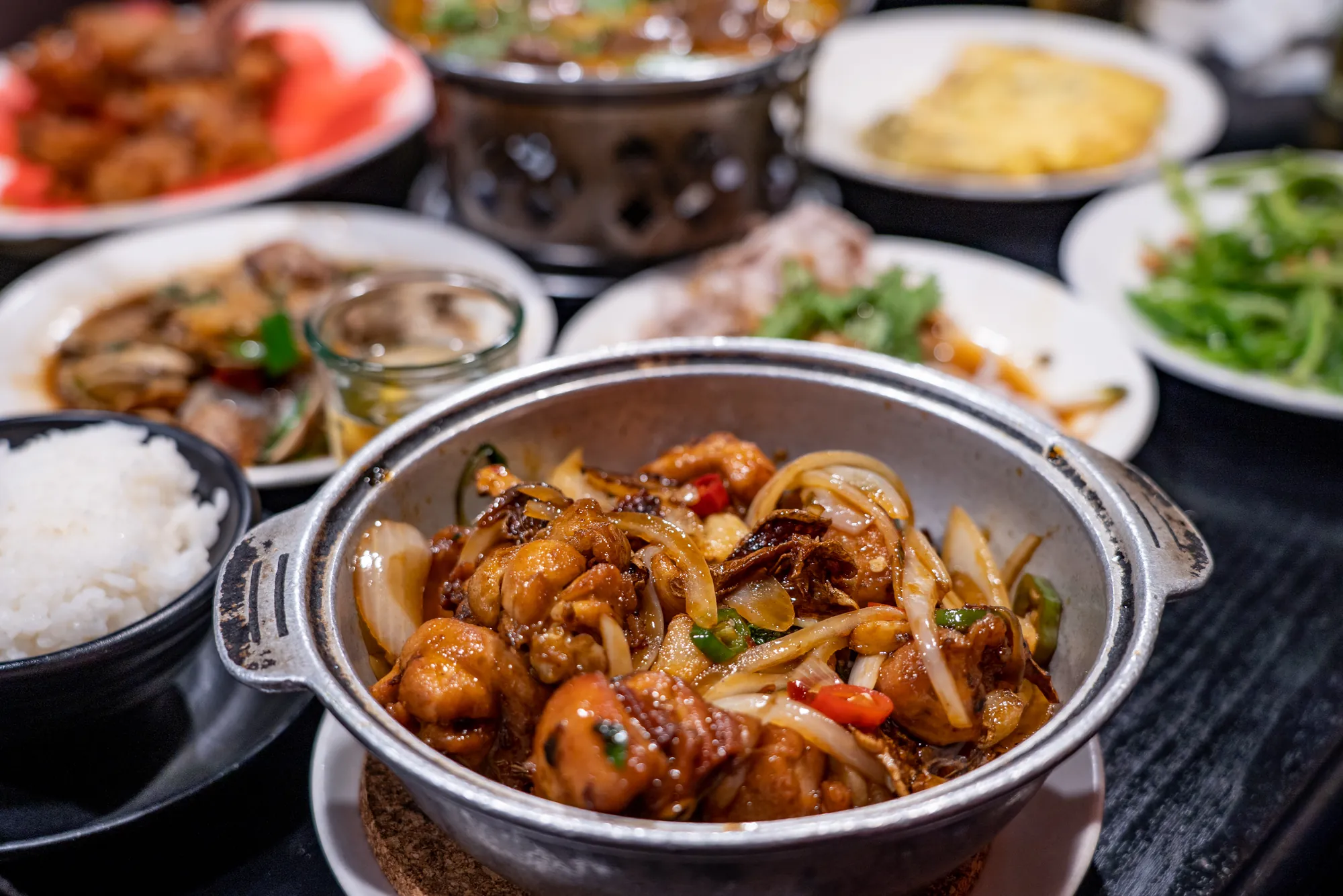 特寫一熱氣騰騰的鐵鍋三杯雞,內有雞肉塊、九層塔、洋蔥和薑片,背景有多道台灣熱炒菜色與一碗白飯。