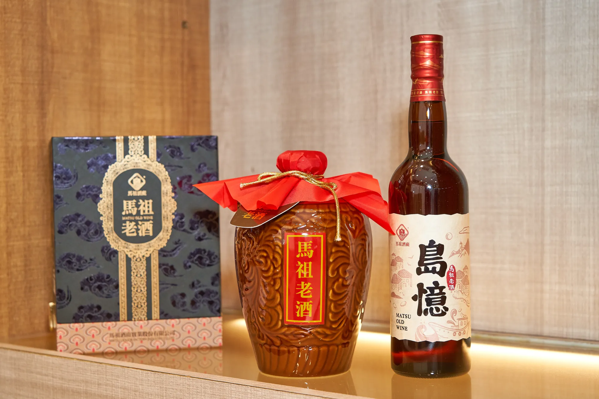 木質展示架上擺放著馬祖老酒系列產品,包含一個藍色禮盒、一個陶甕裝老酒,以及一瓶玻璃瓶裝的『島憶』老酒。