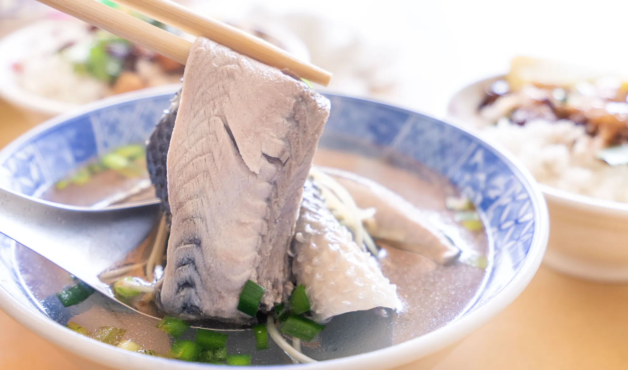用筷子夾起青花瓷碗中的台南虱目魚肚,魚肉肥美鮮嫩,湯頭清澈並撒有翠綠蔥花與薑絲,展現道地台灣小吃風味。