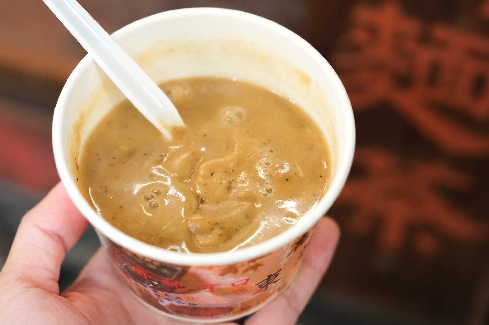 手持一杯濃郁的鹿港傳統美食熱麵茶，杯中插著湯匙，背景隱約可見麵茶攤位的字樣。